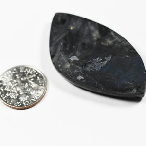 Op de afbeelding: Een marquise-vormige donkergrijze labradoriet edelsteen hanger met zichtbare blauwe iridiscentie en een glad gepolijst oppervlak, geplaatst naast een Amerikaanse dubbeltje om de relatieve grootte van de steen aan te tonen.