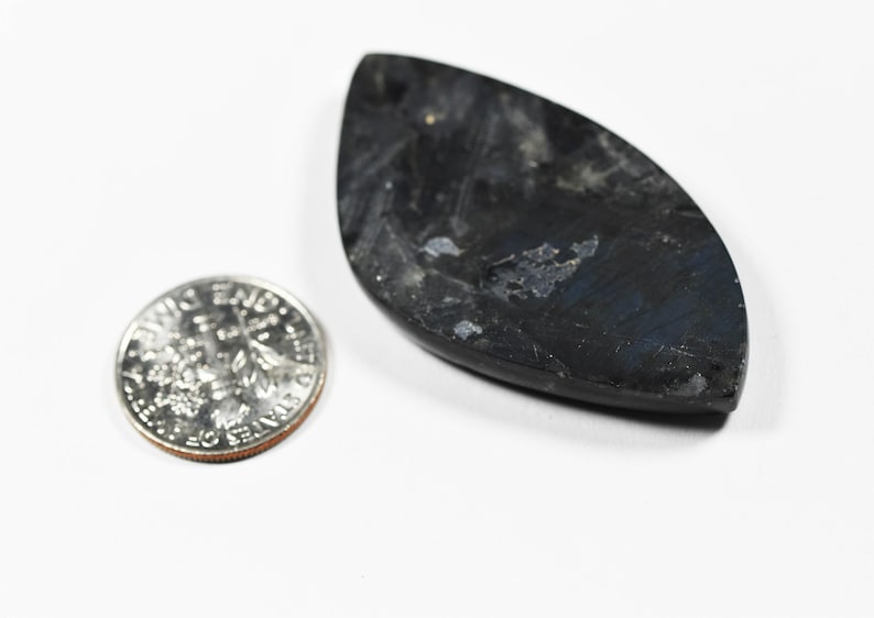 Op de afbeelding: Een marquise-vormige donkergrijze labradoriet edelsteen hanger met zichtbare blauwe iridiscentie en een glad gepolijst oppervlak, geplaatst naast een Amerikaanse dubbeltje om de relatieve grootte van de steen aan te tonen.