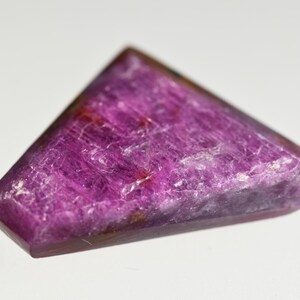 May include: A triangular-shaped gemstone with a vibrant magenta and deep purple hue. The surface of the stone displays a natural, crystalline texture with visible internal inclusions and facets that catch the light, creating a polished appearance.