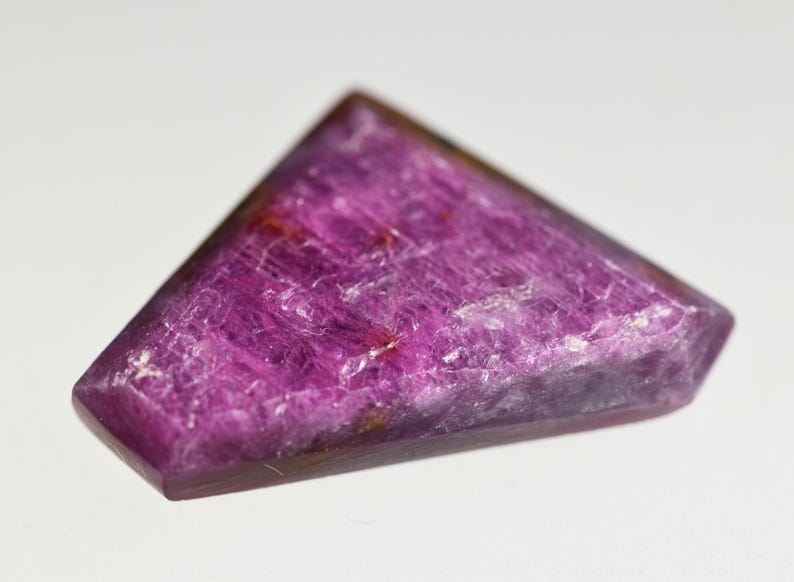 May include: A triangular-shaped gemstone with a vibrant magenta and deep purple hue. The surface of the stone displays a natural, crystalline texture with visible internal inclusions and facets that catch the light, creating a polished appearance.