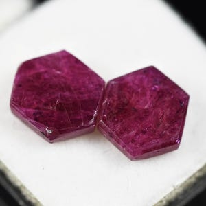 May include: Two loose, hexagonal-cut gemstones with a deep, saturated magenta-red color. The stones feature a flat, polished surface with visible internal inclusions and natural crystalline textures throughout the translucent, vibrant red material.