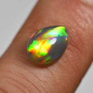 May include: A teardrop-shaped black opal gemstone featuring a smooth, polished surface with vibrant flashes of iridescent green, yellow, and orange color play that shift across the dark, translucent base of the stone.