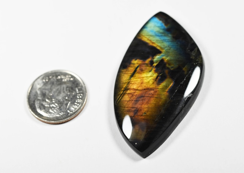 Op de afbeelding: Een marquise-vormige labradoriet edelsteen cabochon met iriserende blauwe en gouden flitsen tegen een donkere basis, geplaatst naast een Amerikaanse kwart dollar munt voor schaal om de relatieve grootte en afmetingen te tonen.