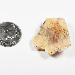 May include: A rough-cut, translucent crystal with orange and black inclusions, next to a U.S. dime for size comparison. The crystal has a light yellow and cream color with a slightly rough texture. The dime is silver in color.