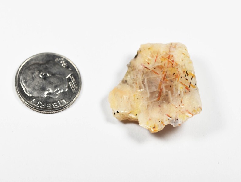 May include: A rough-cut, translucent crystal with orange and black inclusions, next to a U.S. dime for size comparison. The crystal has a light yellow and cream color with a slightly rough texture. The dime is silver in color.
