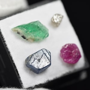 May include: A collection of four raw, uncut gemstones displayed on a white square pad. The set includes a vibrant green emerald, a deep blue sapphire, a bright pink ruby, and a small, translucent, irregular white diamond crystal.