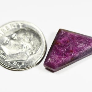 May include: A deep magenta, triangular-shaped gemstone with a faceted surface texture, positioned next to a United States dime coin for scale, which measures approximately 0.705 inches in diameter, providing a clear visual reference for the size.