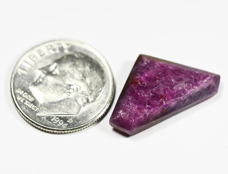 May include: A deep magenta, triangular-shaped gemstone with a faceted surface texture, positioned next to a United States dime coin for scale, which measures approximately 0.705 inches in diameter, providing a clear visual reference for the size.