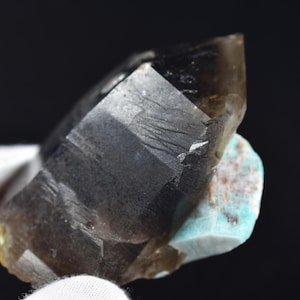 Può includere: Un cristallo di quarzo affumicato con una texture naturale, grezza e ruvida. Il cristallo è di colore marrone scuro e ha un aspetto chiaro e vetroso. Il cristallo è attaccato a un pezzo di amazonite verde.
