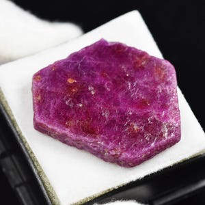 May include: A rough-cut, hexagonal ruby gemstone, showcasing a deep magenta hue with visible texture and natural inclusions. The gem is presented on a white surface within a black display case, highlighting its vibrant color and raw beauty.