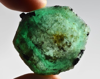Puede incluir: Una esmeralda en bruto, sostenida entre los dedos, que muestra un tono verde vibrante con inclusiones oscuras. La superficie tiene una apariencia texturizada, con áreas de verde más claro y más oscuro. La forma de la gema es irregular, con un aspecto natural y sin pulir.