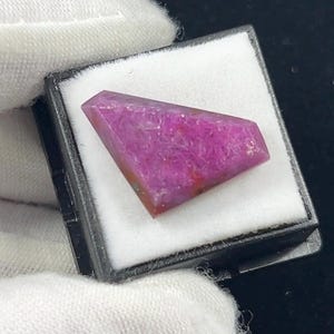 May include: A faceted, trapezoidal-shaped gemstone with a vibrant magenta and pink hue. The stone features a textured, crystalline surface with subtle variations in color intensity, displayed inside a small black box with a white foam insert.