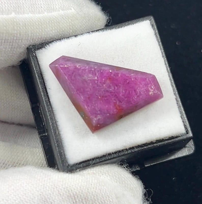 May include: A faceted, trapezoidal-shaped gemstone with a vibrant magenta and pink hue. The stone features a textured, crystalline surface with subtle variations in color intensity, displayed inside a small black box with a white foam insert.