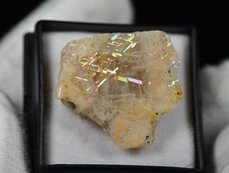 May include: A rough-cut, iridescent sunstone crystal, showcasing a spectrum of colors. The stone is a pale yellow-beige with flashes of rainbow hues. It is presented in a small, black display box with a white interior.