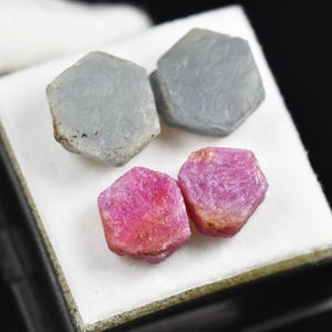 May include: Four rough gemstones, two gray and two pink, are displayed on a white surface within a black box. The gemstones have a hexagonal shape and a natural, unpolished appearance. The pink stones have a vibrant, reddish hue.