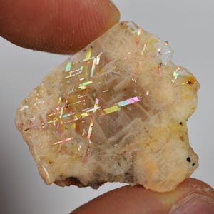 May include: A rough-cut, beige sunstone gemstone with iridescent flashes of color. The stone is held between two fingers, showcasing its natural, irregular shape and the play of light across its surface. The stone has a rough, textured appearance.