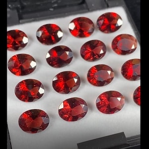 Può includere: Una collezione di 16 pietre preziose ovali di colore rosso. Le pietre preziose sono disposte in file su una superficie bianca.