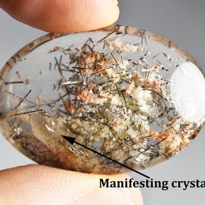 Può includere: Cristallo trasparente di forma ovale con inclusioni interne di minerali neri, arancioni e bianchi. Il cristallo è tenuto tra le dita e presenta la scritta "Manifesting crystal!". Una gemma naturale unica.