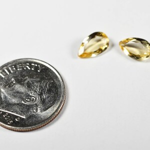 May include: Two pear-shaped, yellow gemstones next to a U.S. dime coin. The gemstones have a polished, translucent appearance. The coin shows the profile of a person and the word "LIBERTY". The background is white.