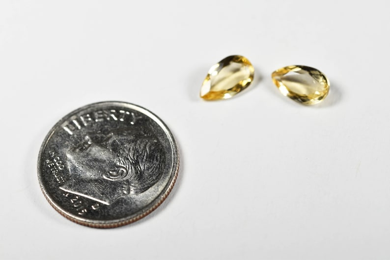 May include: Two pear-shaped, yellow gemstones next to a U.S. dime coin. The gemstones have a polished, translucent appearance. The coin shows the profile of a person and the word "LIBERTY". The background is white.