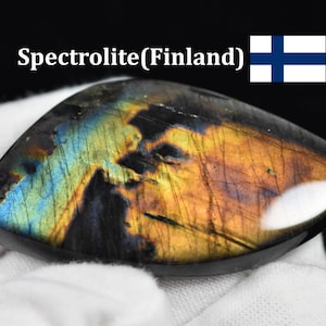 Op de afbeelding: Een gepolijste spectroliet edelsteen met een donkere basis en iriserende blauwe en oranje flitsen. De tekst "Spectrolite(Finland)" staat boven de steen naast een kleine Finse vlag, die een blauw kruis op een witte achtergrond weergeeft.