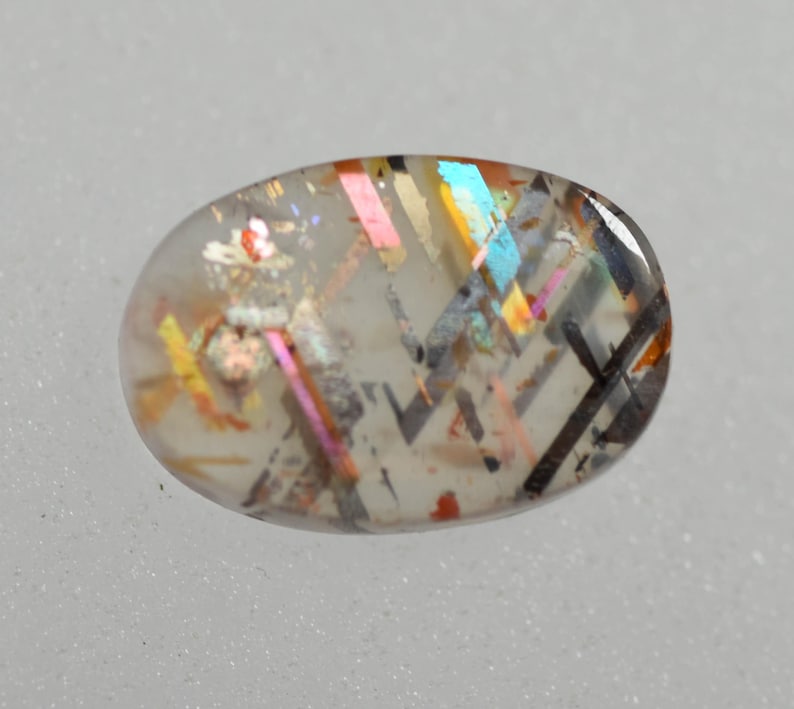 May include: Oval-shaped gemstone with a clear base and internal inclusions. The gemstone features iridescent flashes of color, including pink, blue, and yellow, along with black and brown linear patterns. The stone is set against a light gray background.