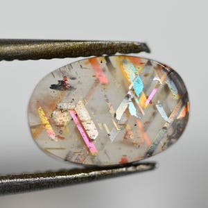 May include: A clear, oval-shaped gemstone with iridescent, colorful inclusions. The gem is held by a pair of tweezers, showcasing its internal patterns of pink, yellow, and blue. The stone's surface is slightly translucent.