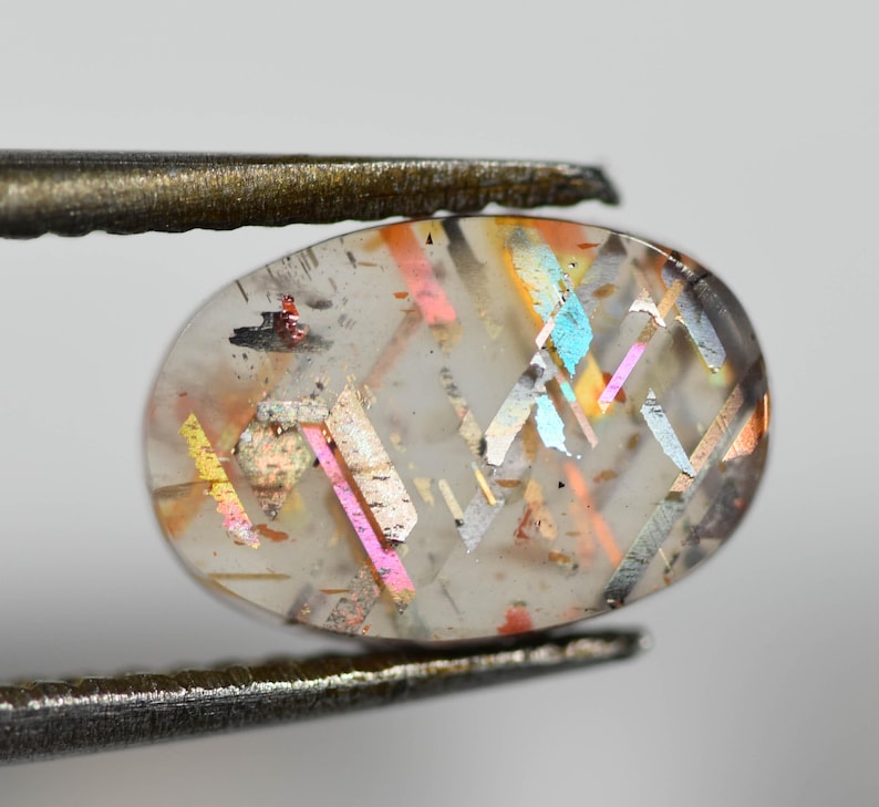 May include: A clear, oval-shaped gemstone with iridescent, colorful inclusions. The gem is held by a pair of tweezers, showcasing its internal patterns of pink, yellow, and blue. The stone's surface is slightly translucent.