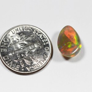 May include: A teardrop-shaped opal gemstone displaying a play-of-color with flashes of red, orange, and green, positioned next to a United States one dime coin for scale to show the relative size of the stone.