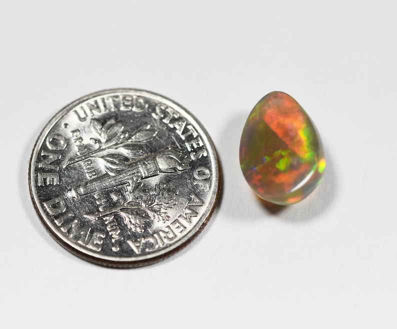 May include: A teardrop-shaped opal gemstone displaying a play-of-color with flashes of red, orange, and green, positioned next to a United States one dime coin for scale to show the relative size of the stone.