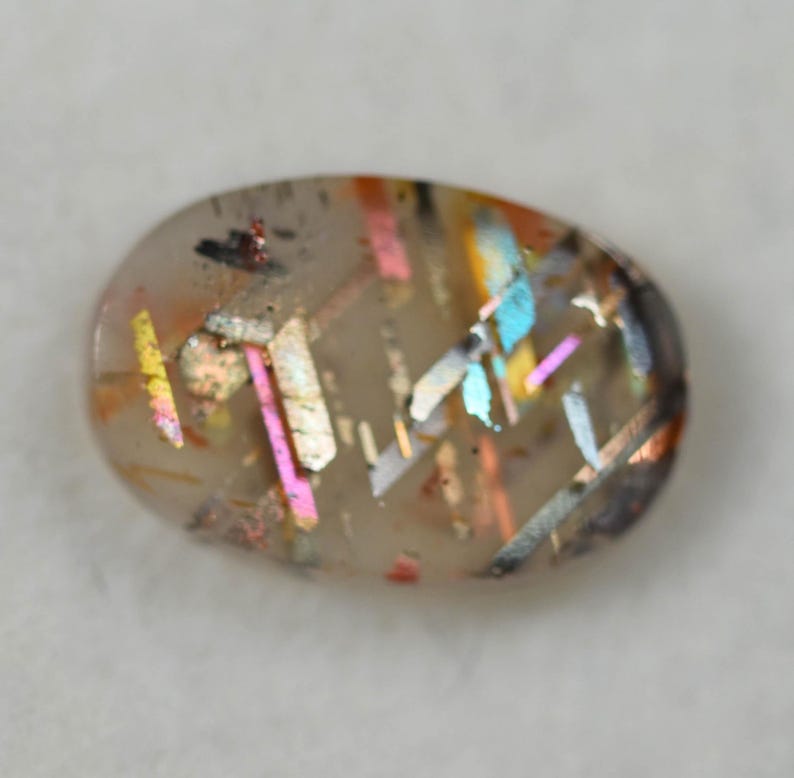 May include: An oval-shaped gemstone with a translucent base, showcasing internal inclusions of various colors. The inclusions appear as iridescent streaks of pink, blue, yellow, and orange, creating a unique, eye-catching pattern. The gemstone has a polished surface.