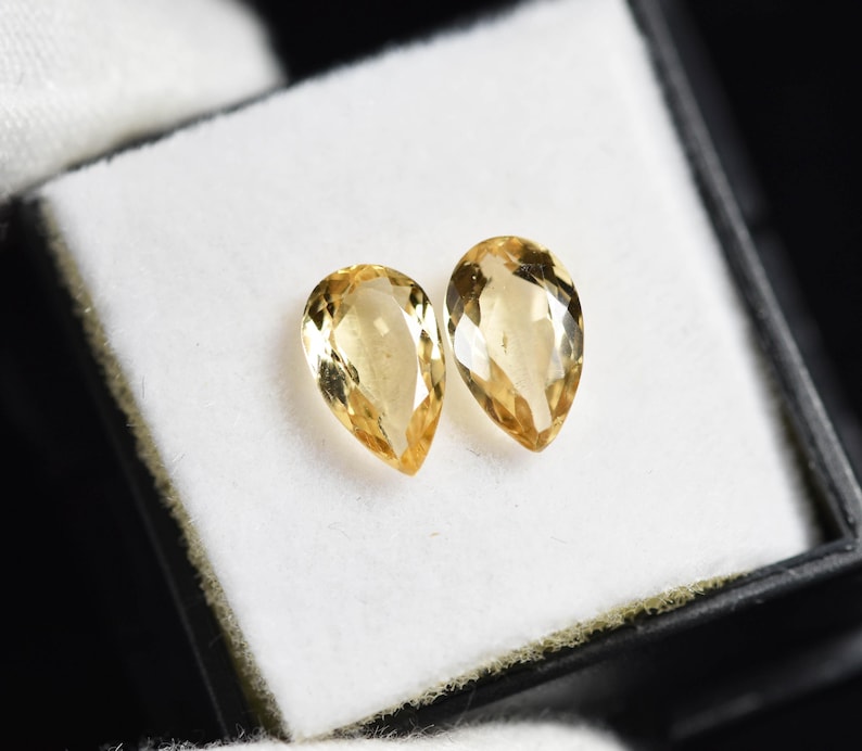 May include: Two pear-shaped, faceted gemstones with a warm, golden-yellow hue. The gems are displayed in a white box with a black border, creating a striking contrast. The stones are likely for jewelry making.
