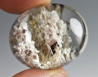 Op de afbeelding: Een gepolijste, ovaalvormige, heldere kwarts-edelsteen met witte en zilveren minerale insluitingen. De steen heeft een glad, afgerond oppervlak met een transparant uiterlijk en toont een textuurrijk, kristallijn cluster aan de binnenkant.