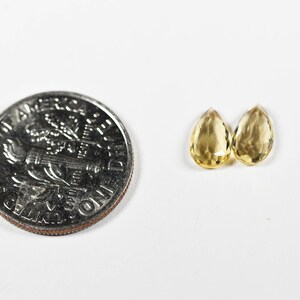 May include: Two faceted, teardrop-shaped, yellow gemstones next to a U.S. dime. The gemstones are a light yellow color and appear to be transparent. The dime is silver and shows the profile of a person.