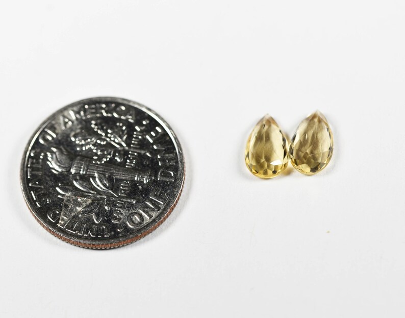 May include: Two faceted, teardrop-shaped, yellow gemstones next to a U.S. dime. The gemstones are a light yellow color and appear to be transparent. The dime is silver and shows the profile of a person.