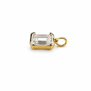 May include: A gold pendant featuring a rectangular emerald-cut clear gemstone set in a polished gold bezel frame. The pendant includes a small circular gold jump ring attached to one side, designed for easy connection to a chain or necklace.