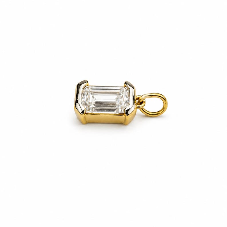 May include: A gold pendant featuring a rectangular emerald-cut clear gemstone set in a polished gold bezel frame. The pendant includes a small circular gold jump ring attached to one side, designed for easy connection to a chain or necklace.