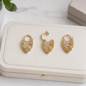 May include: Three gold-colored padlocks with a heart-shaped design, each adorned with sparkling crystals. One padlock is open, revealing its intricate mechanism. The padlocks are displayed on a white rectangular box.