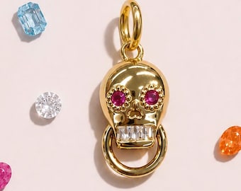 May include: A gold-toned skull pendant featuring two round pink gemstone eyes and a row of rectangular white baguette-cut teeth. A circular gold ring hangs from the jaw, and a gold jump ring is attached to the top of the skull for hanging.