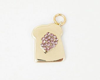 May include: A gold-colored charm in the shape of a slice of bread, with a cluster of pink crystals representing jelly. The charm has a gold ring at the top for attachment. The design is simple and elegant.