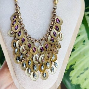 May include: A gold-toned statement necklace with a cascade of circular pendants. Each pendant is adorned with a teardrop-shaped, faceted gem in shades of purple, clear, and amber. The necklace is displayed on a cream-colored bust.