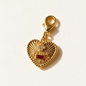 May include: A gold-toned heart-shaped charm featuring a lobster clasp, a central red rectangular stone, a small cross with clear stones, and the embossed text OUCHIER. The surface has a sunburst pattern and a beaded border.