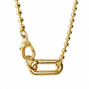 May include: A gold-toned ball chain necklace featuring a lobster claw clasp with a small embedded clear stone. The clasp is attached to a large, elongated oval link, creating a prominent focal point for the jewelry piece against a plain background.