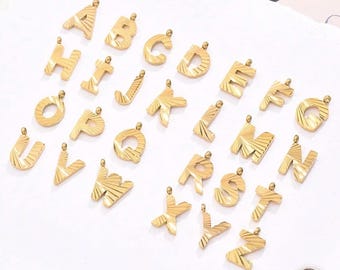 May include: Gold-colored alphabet pendants, each letter with a textured, sunburst design. The pendants are arranged on a white surface, showcasing the full alphabet from A to Z. Each letter has a small loop at the top for attaching to a chain.