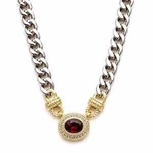 May include: A two-tone necklace featuring a thick silver-colored curb chain connected to a central gold-colored oval pendant. The pendant contains a deep red oval gemstone surrounded by a halo of small clear stones and textured gold rope details.