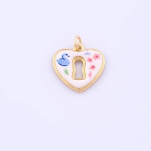 May include: A heart-shaped pendant with a gold-colored border and bail. The pendant features a white enamel surface with a keyhole cutout. A blue swan and pink flowers decorate the surface. The pendant is likely for jewelry making.