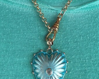 May include: A gold-toned chain necklace with a heart-shaped pendant. The pendant is a light blue color with a textured design and a small, clear stone in the center. The necklace is displayed against a teal background.