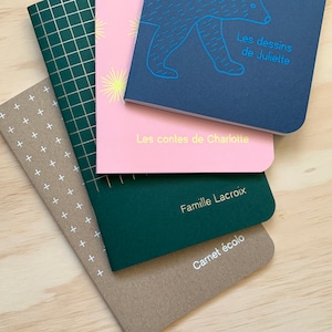 Peut inclure: Une pile de quatre cahiers avec des couvertures de couleurs différentes. Le cahier du dessus est bleu avec une illustration d'un ours blanc et le texte "Les dessins de Juliette". Le deuxième cahier est rose avec le texte "Les contes de Charlotte". Le troisième cahier est vert avec le texte "Famille Lacroix". Le cahier du dessous est marron avec le texte "Carnet écolos".