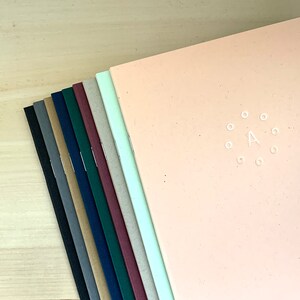 K&ouml;nnte beinhalten: Ein Stapel von zehn Notizb&uuml;chern mit verschiedenen farbigen Umschl&auml;gen. Das oberste Notizbuch ist pfirsichfarben und hat einen wei&szlig;en Kreis mit dem Buchstaben A in der Mitte.