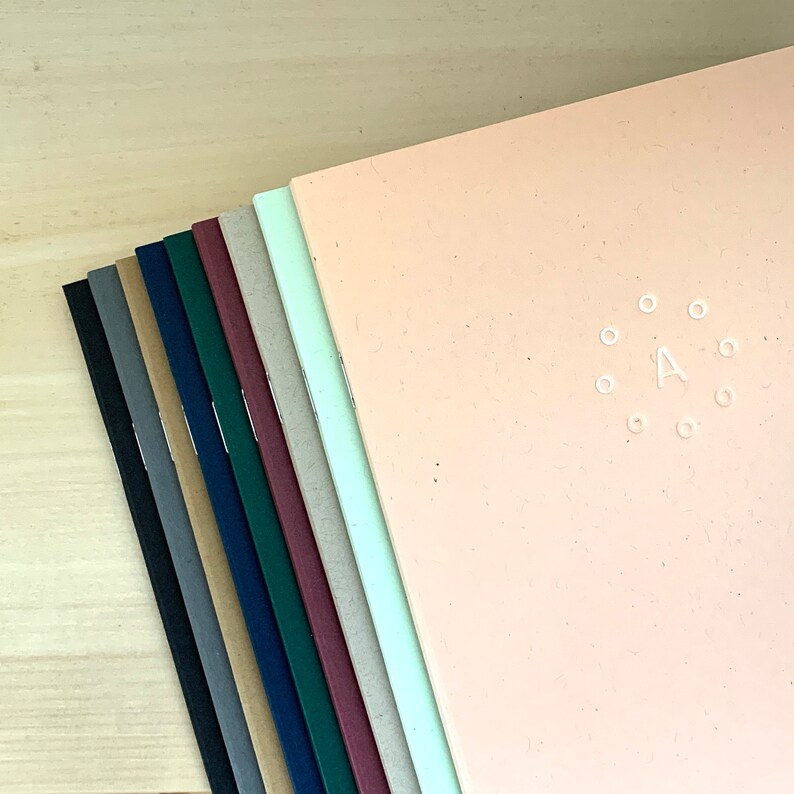 K&ouml;nnte beinhalten: Ein Stapel von zehn Notizb&uuml;chern mit verschiedenen farbigen Umschl&auml;gen. Das oberste Notizbuch ist pfirsichfarben und hat einen wei&szlig;en Kreis mit dem Buchstaben A in der Mitte.